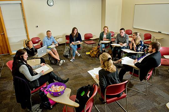 A female professor sitting in a round classroom configuration with several students discussing public relations