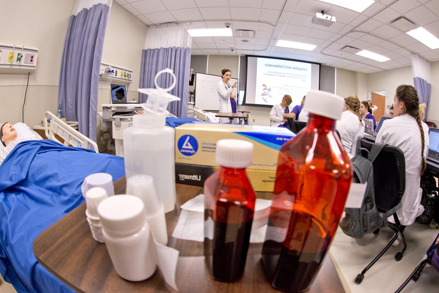 在护理模拟实验室里，穿着白大褂的学生们在投影仪前听女教授讲课，周围是医疗设备和处方瓶