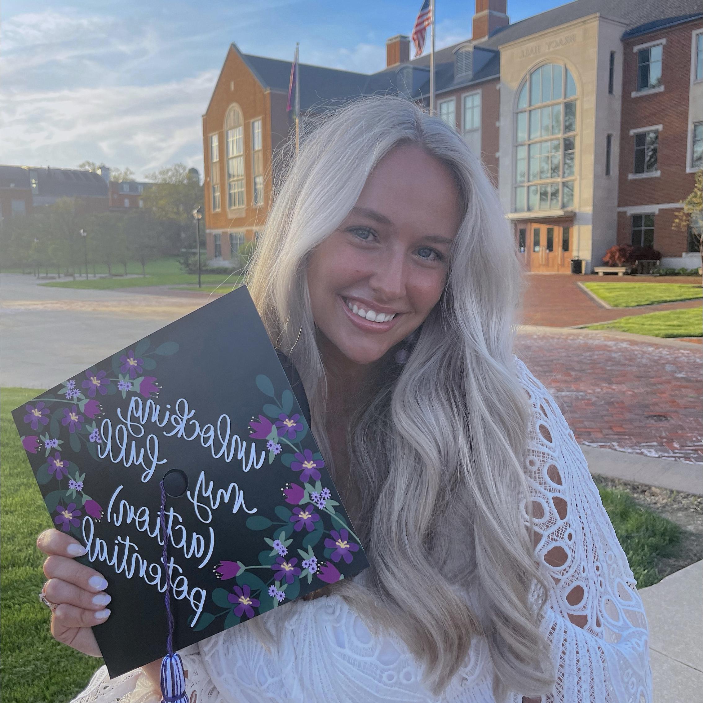Katie Wilson stands in front of Bracy Hall with a decorated graduation cap with purple flowers. The hat says in white calligraphy-style writing, “unlocking my full (action) potential”
